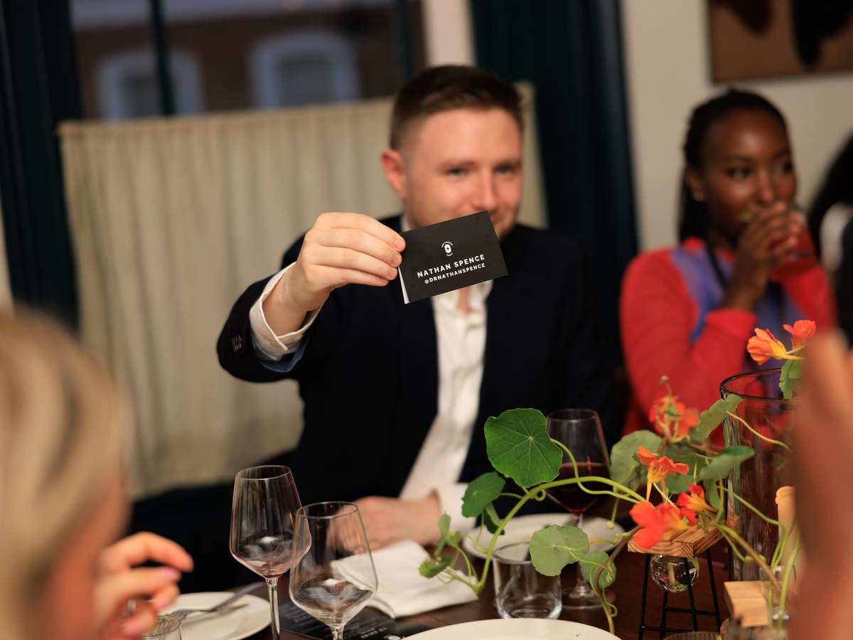 Nathan Spence at a FIGS-related event holding a business card while seated at a dinner